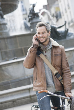 handsome man at phone on a bicycle with fountain in background