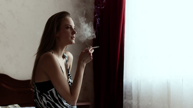 Girl in shirt sitting on a bed near the window smokes