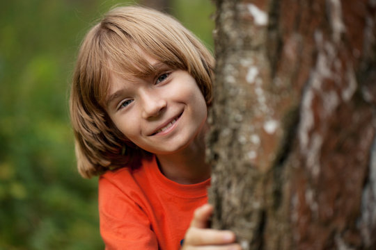 Boy in red T-shirt peeking out from behind a tree trunk