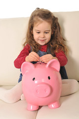 Cute little girl putting coin in huge piggy bank on sofa