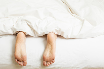 Woman's Feet Sticking Out Of Blanket On Bed At Home