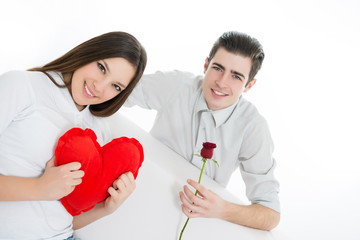 Beautiful,young woman with a heart shape pillow ,man standing behind her and holding a red rose flower, looking at camera