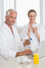 Smiling couple using laptop at breakfast in bathrobes