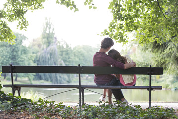 Fototapeta premium Rear view of romantic young couple sitting on bench at lakeside