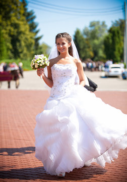 Smiling brunette bride in fluffy dress walking at park alone