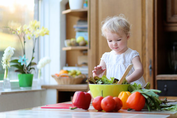 Obraz na płótnie Canvas Funny toddler girl mixing vegetables in the bowl preparing salad