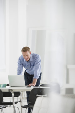Business. A Man Standing Over A Desk, Leaning Down To Use A Laptop Computer. 