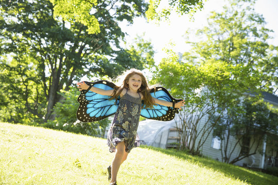  A Child Running Across The Lawn In Front Of A Farmhouse, Wearing Large Irridescent Blue Butterfly Wings And With Her Arms Outstretched. 