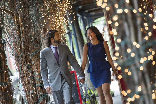 Business People. Two People, A Man And Woman Holding Hands And Walking Under A Pergola, Lit With Fairy Lights. 