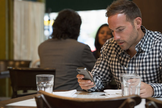 Business People. Two People Talking To Each Other, And A Man At A Separate Table Checking His Phone. 