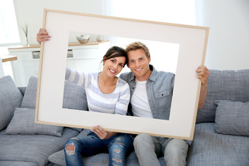 Cheerful young couple holding picture frame to look through - obrazy, fototapety, plakaty
