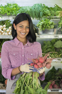 A farm stand with rows of freshly picked vegetables for sale. A woman holding a bunch of carrots.