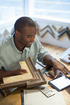 A young man at his workbench in a picture framing studio. Surrounded by samples. Using a digital tablet.