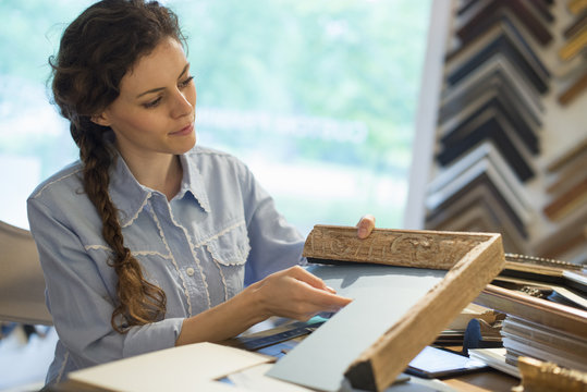 A woman in a picture framer's workshop, choosing frames and cardboard mounts. Surrounded by samples.