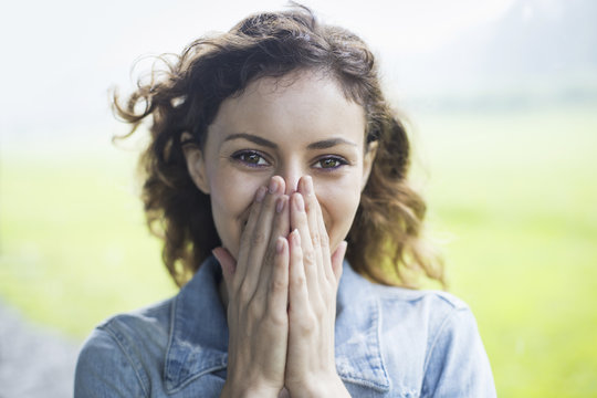 A young woman in a rural landscape, with windblown curly hair. Covering her face with her hands, and laughing.
