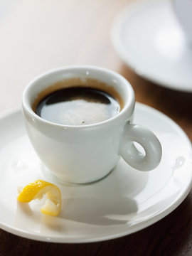 A cup of black coffee in a white china cup with a small twist of lemon peel in the saucer.
