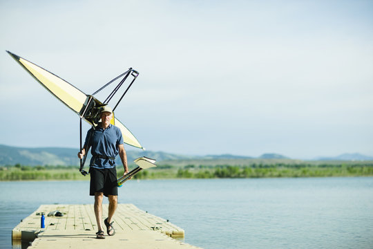 A middle-aged man carrying oars and a rowing shell on his shoulders, on a pontoon.