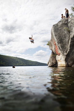 A group of young people jumping from a height from a cliff into the still waters of a lake. 