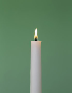 A thin white wax candle with a small  lit flame with a green background.