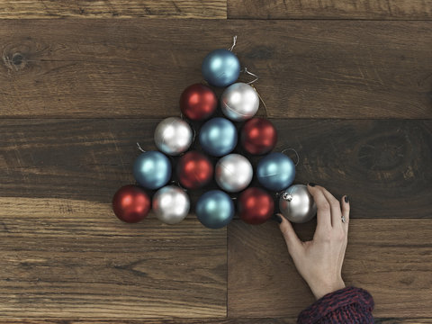 A collection of blue, red and silver ornaments arranged in a triangular shape on a wooden board. A Christmas tree shape. A person's hand placing the final ball.