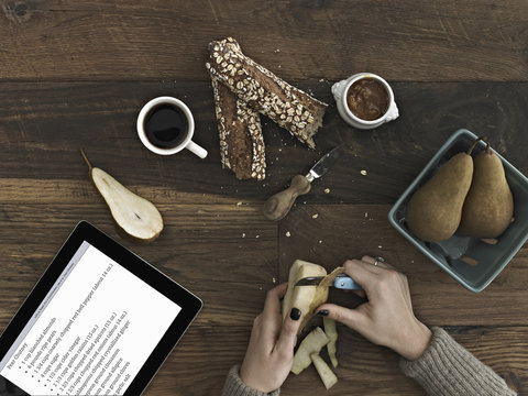 A person using a peeling knife to peel a pear. A computer tablet device with the screen displaying instructions, a recipe.