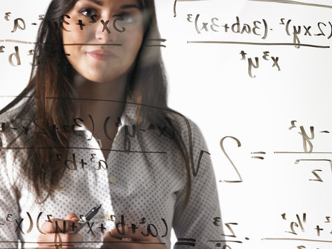 A young woman looking at a mathematical equation written out with black marker on a clear seethrough wall.