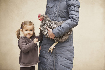A woman in a grey coat holding a black and white chicken with a red coxcomb under one arm. A young girl beside her holding her other hand