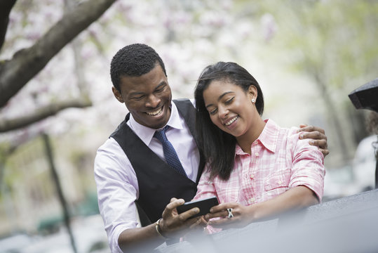 City life in spring. Young people outdoors in a city park. A couple side by side, with his arm around her shoulders, looking at a smart phone and smiling.