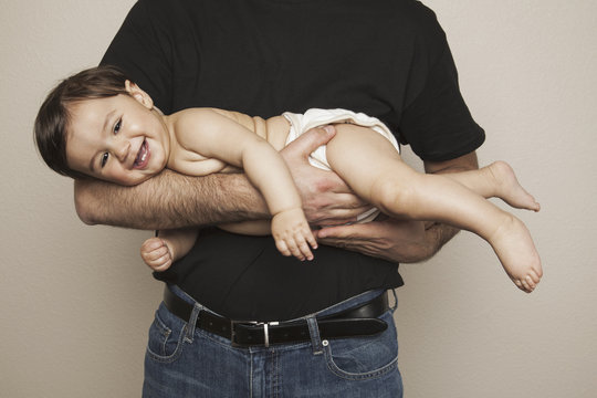 A young boy wearing cloth diapers being held by his father in his arms.