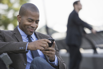 Business people in the city. Keeping in touch on the move. A man seated using his smart phone. A man in the background.
