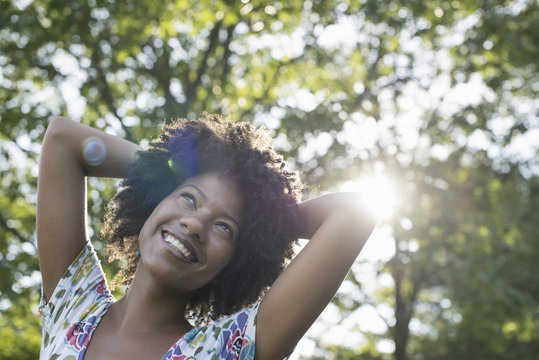 A young woman in a flowered summer dress with her hands behind her head, smiling and looking up.