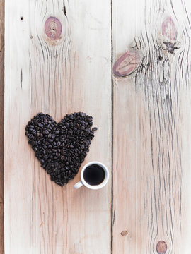 Overhead view of a heap of roasted organic coffee beans in a heart shape, arranged on a tabletop. A cup of black coffee.
