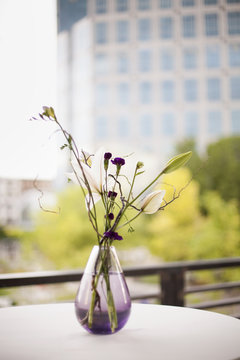 A table on a terrace in the city. A vase of flowers. Small purple flowers, and white lily and orchid blooms. 