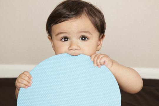 A young 8 month old baby boy wearing cloth diapers, holding a large blue disc and chewing the edge.