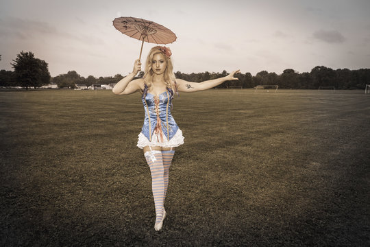 A young woman wearing a laced basque and frilly bloomers, striped stockings and suspenders. Holding a paper sunshade umbrella. A carnival circus outfit. 
