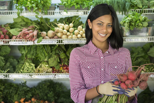 A farm stand with rows of freshly picked vegetables for sale. A woman holding a bunch of carrots.