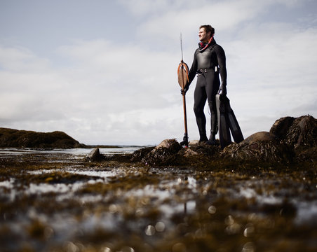 A man in a wetsuit, standing on the shore with a large spear fishing harpoon.