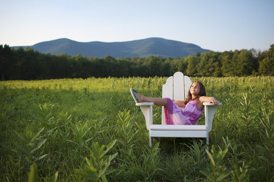 A young girl sitting in a traditional wooden Adirondack style chair in a field at evening light. 