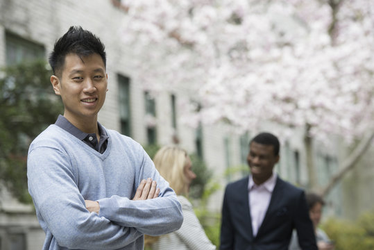 City life in spring. Young people outdoors in a city park. A man with his arms folded and two people talking in the background under the trees in blossom.