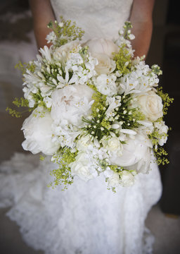 A woman in a white dress, a bride holding a bridal bouquet of white flowers, large white roses and peonies, with delicate yellow flowers and green leaves.  