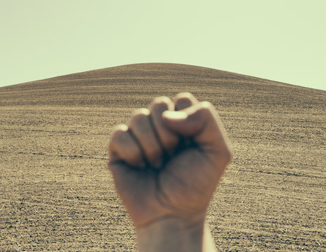 A hand bunched up and making a fist, in a gesture of defiance or determination, against the background of a ploughed field and farmland. 