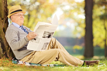 Smiling senior gentleman on grass reading a newspaper in park at
