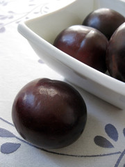 Black plums in a square white bowl.