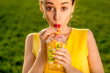 Young woman drinking fruits from the transparent cup on green gr