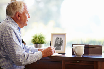 Senior Man Writing Memoirs In Book Sitting At Desk - obrazy, fototapety, plakaty