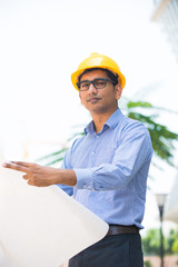 indian male architect holding plans with construction background