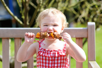 Funny toddler girl eating bbq meat outdoors in the garden - obrazy, fototapety, plakaty
