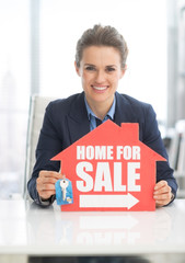 Happy realtor woman showing keys and home for sale sign