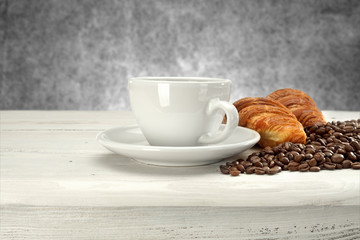 A white cup of hot coffee and croissants and scattered coffee beans on a table with a light gray...