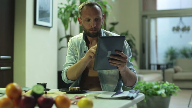 man working on a tablet and eating sandwich at home